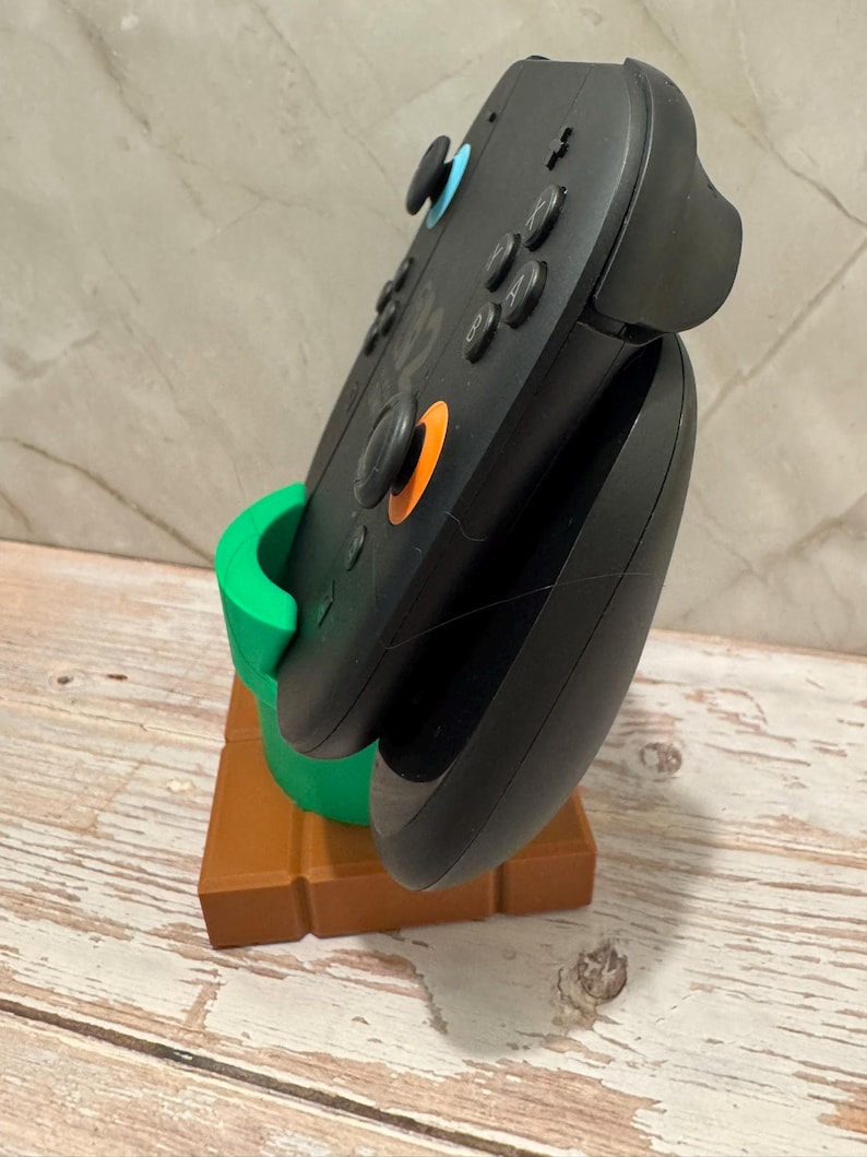 May include: Black video game controller in a green and brown stand. The stand is designed to resemble a green pipe on a brown brick base. The controller features blue and orange buttons.