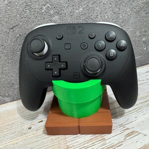 K&ouml;nnte beinhalten: Schwarzer Nintendo Switch Pro Controller auf einem gr&uuml;nen Rohr und einem braunen, ziegelartigen St&auml;nder. Der Controller hat wei&szlig;e Akzente und verschiedene Tasten. Der St&auml;nder ist ein 3D-Druck-Design.