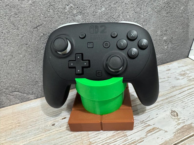 K&ouml;nnte beinhalten: Schwarzer Nintendo Switch Pro Controller auf einem gr&uuml;nen Rohr und einem braunen, ziegelartigen St&auml;nder. Der Controller hat wei&szlig;e Akzente und verschiedene Tasten. Der St&auml;nder ist ein 3D-Druck-Design.