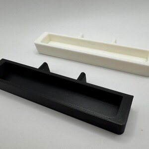 May include: Two rectangular trays, one black and one white, are displayed against a white background. The black tray is in the foreground, with a recessed interior and two triangular protrusions. The white tray is in the background, with a similar design.