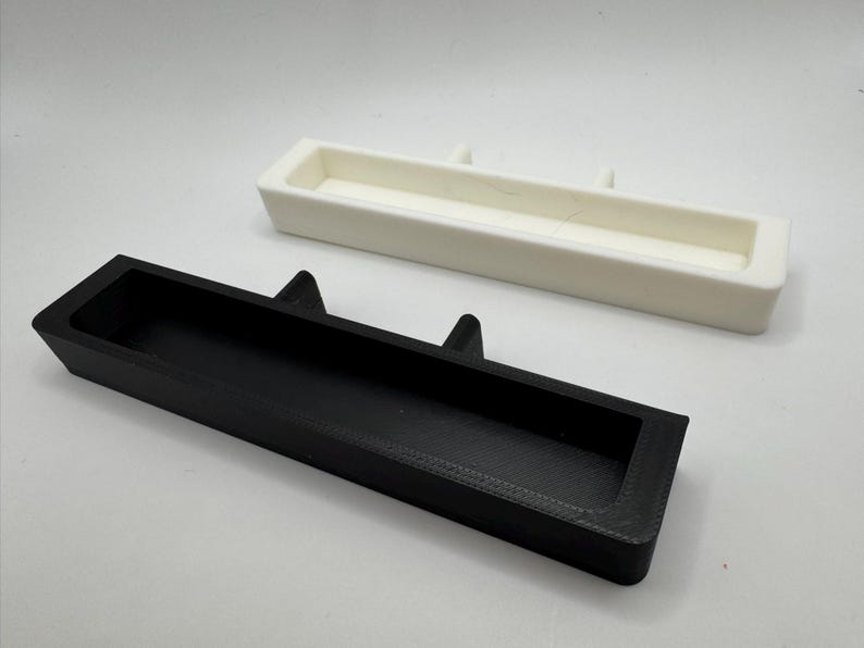 May include: Two rectangular trays, one black and one white, are displayed against a white background. The black tray is in the foreground, with a recessed interior and two triangular protrusions. The white tray is in the background, with a similar design.