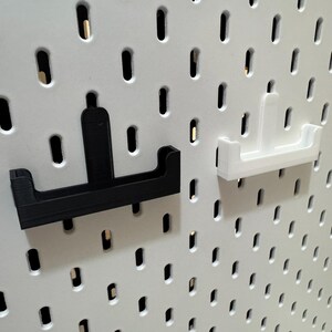 May include: Two pegboard hooks, one black and one white, are displayed against a white pegboard. The hooks have a U-shaped design with a vertical support. The pegboard has evenly spaced oval holes.