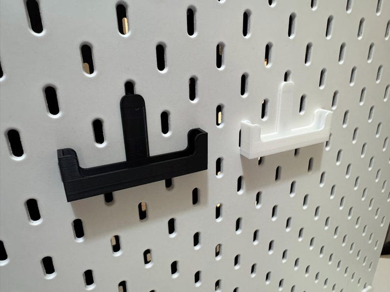 May include: Two pegboard hooks, one black and one white, are displayed against a white pegboard. The hooks have a U-shaped design with a vertical support. The pegboard has evenly spaced oval holes.