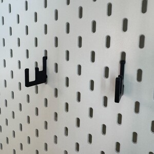 May include: White pegboard with evenly spaced oval holes. Two black hooks are inserted into the board. The hooks are designed to hold items. The pegboard is a versatile storage and organization solution for various spaces.