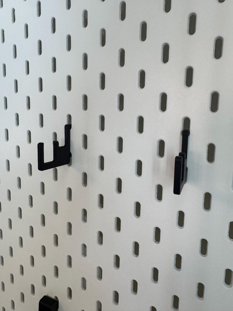 May include: White pegboard with evenly spaced oval holes. Two black hooks are inserted into the board. The hooks are designed to hold items. The pegboard is a versatile storage and organization solution for various spaces.