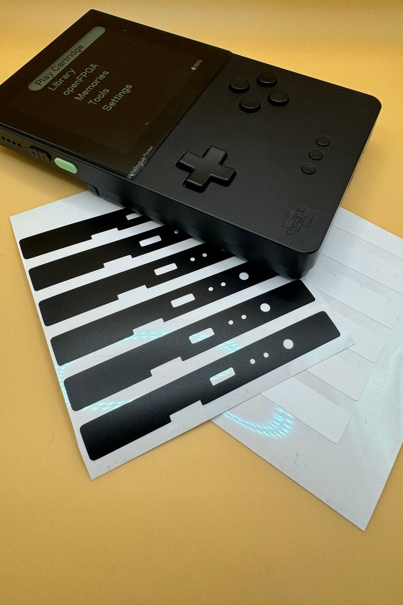 May include: A black handheld gaming console with a screen displaying the text "Play Cartridge", "Library", "openFPcGA", "Memories", "Tools", and "Settings". The console has a directional pad, four buttons, and a green button on the side. The console is on top of a sheet of white paper with black and white stickers.