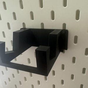 K&ouml;nnte beinhalten: Schwarze Wandhalterung aus Kunststoff mit zwei Haken. Die Halterung ist f&uuml;r Gegenst&auml;nde wie Kopfh&ouml;rer oder Kabel konzipiert.