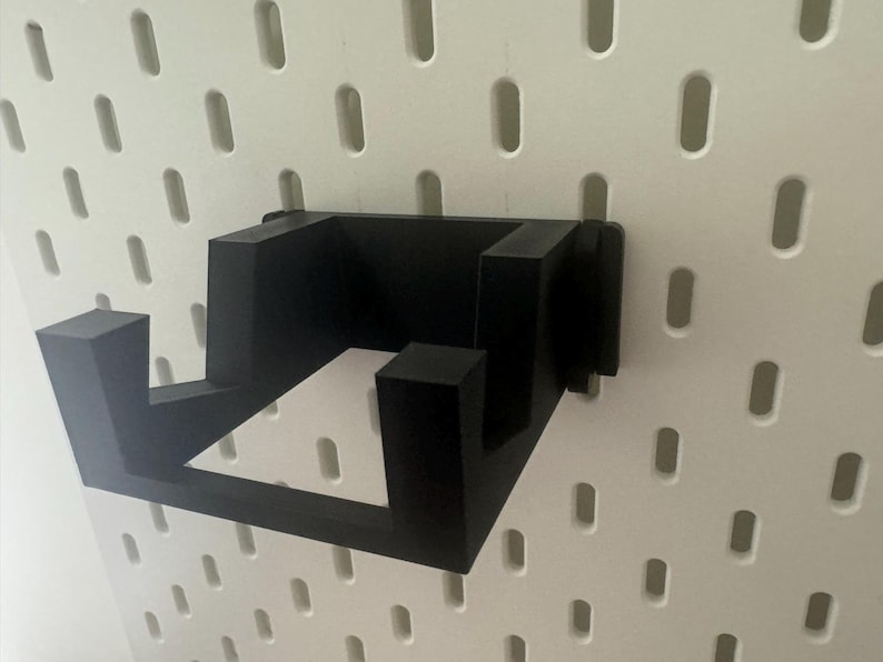 K&ouml;nnte beinhalten: Schwarze Wandhalterung aus Kunststoff mit zwei Haken. Die Halterung ist f&uuml;r Gegenst&auml;nde wie Kopfh&ouml;rer oder Kabel konzipiert.