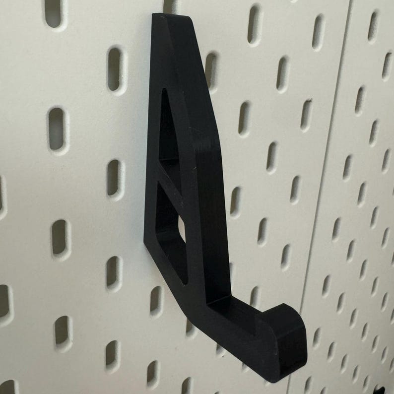 May include: Black plastic wall hook with a triangular shape and a hook on the bottom. The hook is mounted on a white pegboard.