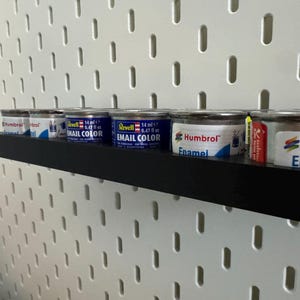 May include: A black shelf holds several small paint cans against a white pegboard. The cans are silver with blue and white labels, some labeled "Revell Email Color" and "Humbrol Enamel".