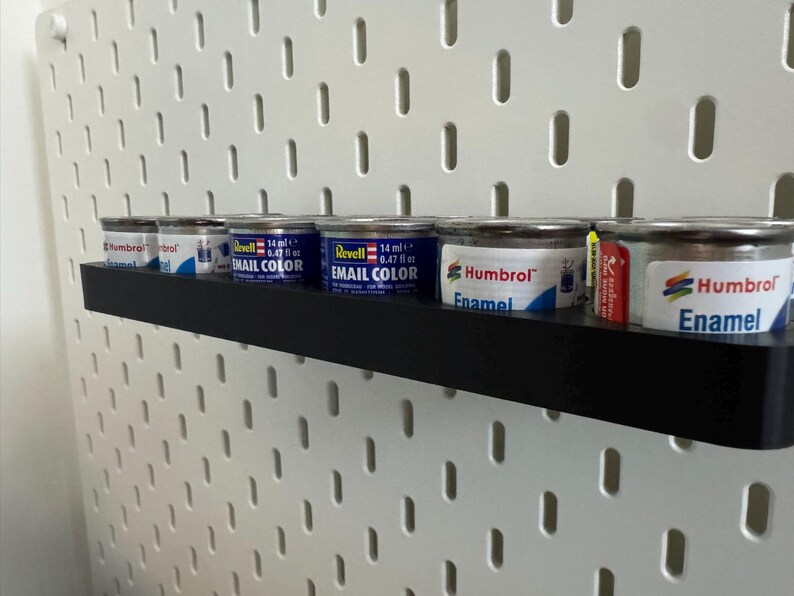 May include: A black shelf holds several small paint cans against a white pegboard. The cans are silver with blue and white labels, some labeled "Revell Email Color" and "Humbrol Enamel".