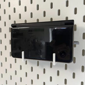 Puede incluir: Una consola de juegos portátil negra está montada en una pared perforada blanca con soportes blancos. La consola está cerrada y refleja el entorno. La pared perforada tiene agujeros ovalados espaciados.