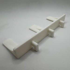 May include: A white plastic component with a rectangular body and several protruding tabs. The object has a clean, minimalist design and appears to be a structural part or connector. The item is designed to fit into a larger structure.