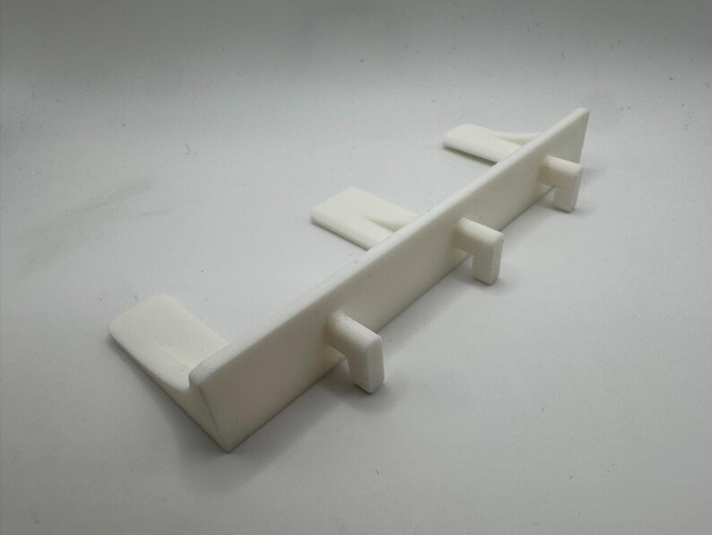 May include: A white plastic component with a rectangular body and several protruding tabs. The object has a clean, minimalist design and appears to be a structural part or connector. The item is designed to fit into a larger structure.