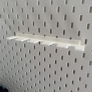 May include: A white plastic shelf designed for a pegboard, featuring multiple slots. The shelf is mounted on a white pegboard with evenly spaced holes. It's a practical storage solution for small items, measured in centimetres.