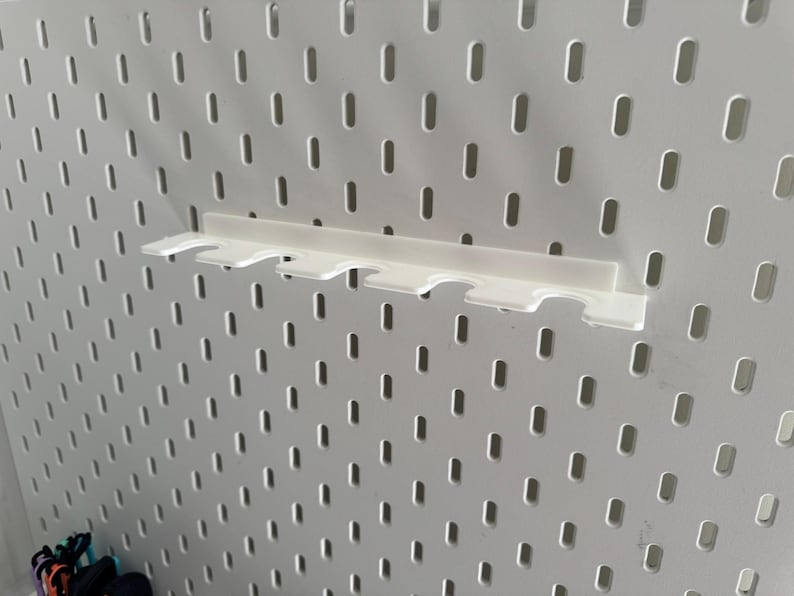 May include: A white plastic shelf designed for a pegboard, featuring multiple slots. The shelf is mounted on a white pegboard with evenly spaced holes. It's a practical storage solution for small items, measured in centimetres.