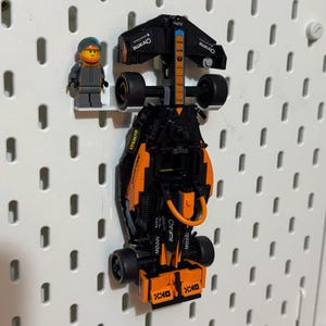 May include: A black and orange toy racing car with the word "Chrome" on the side. The car is hanging on a pegboard wall. A small Lego figure of a racing driver is standing next to the car.