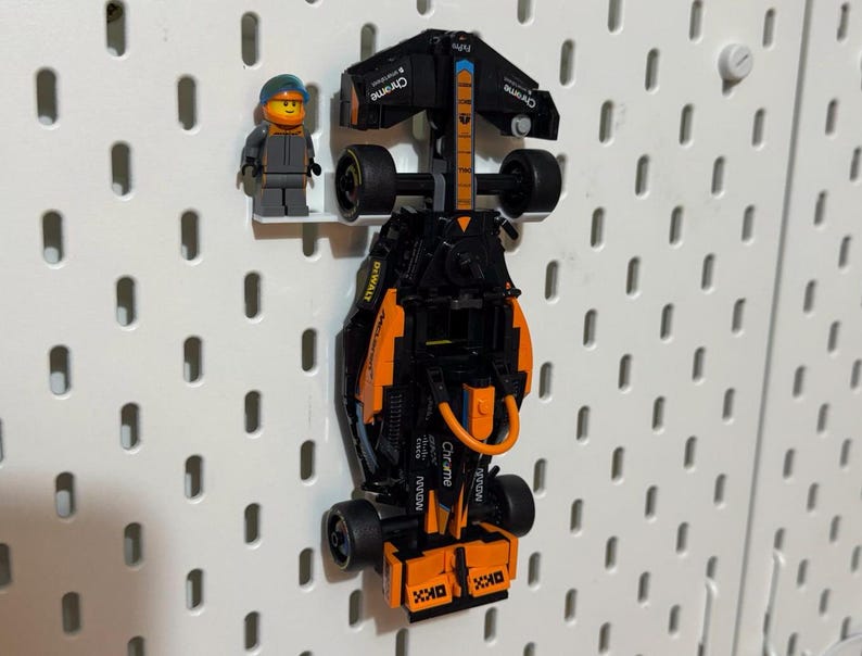 May include: A black and orange toy racing car with the word "Chrome" on the side. The car is hanging on a pegboard wall. A small Lego figure of a racing driver is standing next to the car.