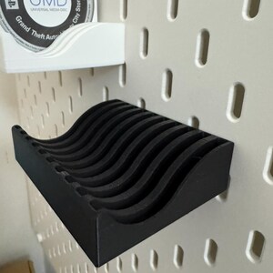 以下が含まれることがあります： 黒い波型のメディアディスク用棚。白いペグボードに取り付けられています。上には、"Grand Theft Auto City Stories"とラベルされたUMDディスクを保持する白い棚があります。