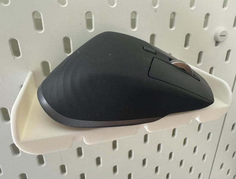 May include: A white plastic mouse holder with a black wireless mouse resting on top. The holder is designed to be mounted on a pegboard wall.
