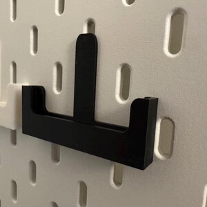 Op de afbeelding: Zwarte pegboardhaak met een verticale steun en een horizontale basis, ontworpen om in de gaten van een pegboard te passen. Er is ook een witte pegboardhaak zichtbaar. De haken zijn gemaakt van een stevig materiaal.
