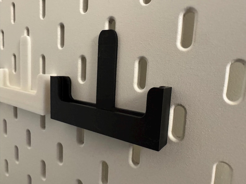 Op de afbeelding: Zwarte pegboardhaak met een verticale steun en een horizontale basis, ontworpen om in de gaten van een pegboard te passen. Er is ook een witte pegboardhaak zichtbaar. De haken zijn gemaakt van een stevig materiaal.