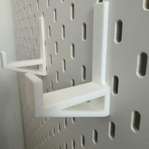 May include: White pegboard shelf brackets. The brackets are L-shaped and designed to fit into the holes of a white pegboard. The brackets are made of a solid, opaque white material. The pegboard has evenly spaced, vertical slots.