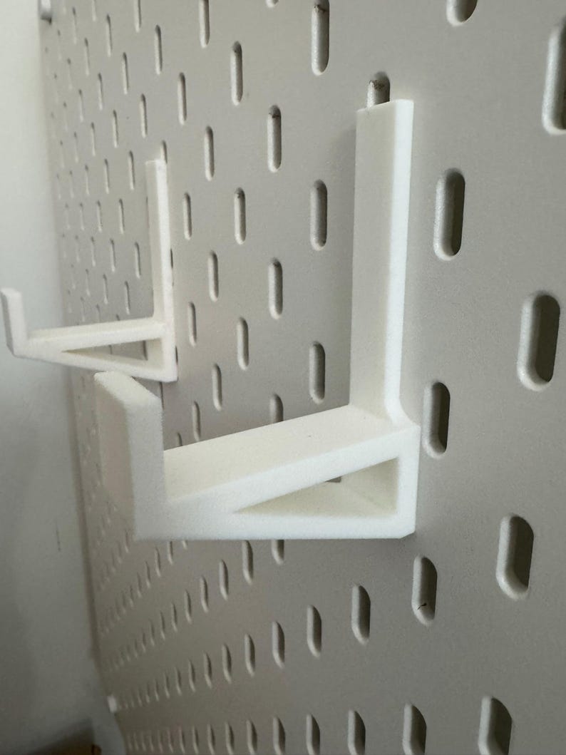 May include: White pegboard shelf brackets. The brackets are L-shaped and designed to fit into the holes of a white pegboard. The brackets are made of a solid, opaque white material. The pegboard has evenly spaced, vertical slots.