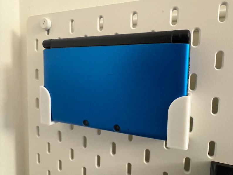 Puede incluir: Una consola de juegos port&aacute;til azul est&aacute; montada en una pared perforada blanca con un soporte blanco. La consola tiene una parte superior negra y est&aacute; asegurada por dos soportes blancos. La pared perforada tiene una serie de agujeros ovalados.