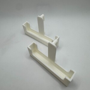 May include: Two white, rectangular 3D-printed objects. Each has a trough-like shape with a raised, vertical support in the center. The objects are designed to hold or support something, possibly for a shelf or display.
