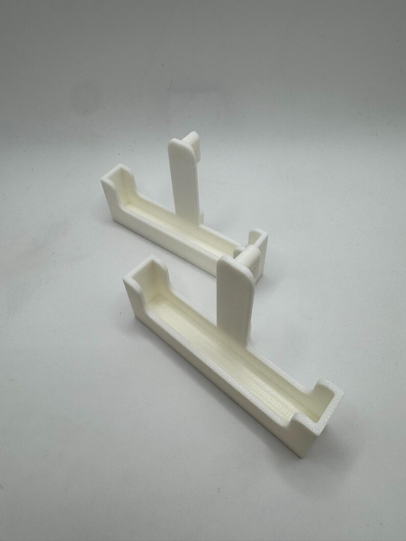 May include: Two white, rectangular 3D-printed objects. Each has a trough-like shape with a raised, vertical support in the center. The objects are designed to hold or support something, possibly for a shelf or display.