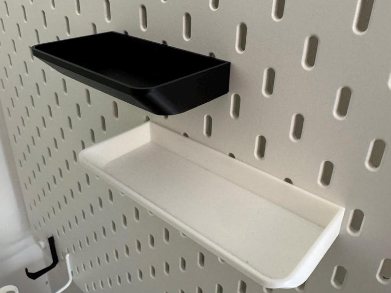 Puede incluir: Dos estantes de pared, uno negro y otro blanco, est&aacute;n montados en una pared de tablero perforado blanco.