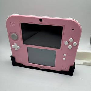May include: A pink Nintendo 2DS handheld game console with a black stand. The console features a top and bottom screen, a directional pad, and various buttons. The stand is black and holds the console upright.