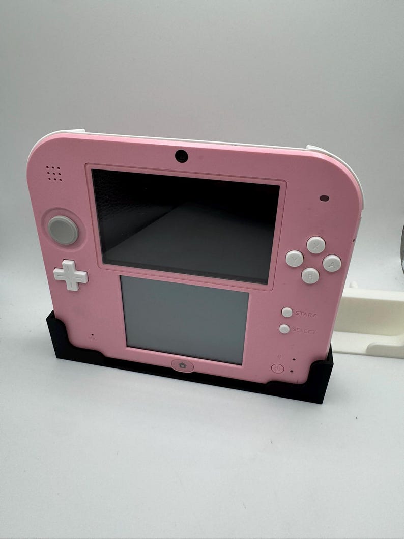 May include: A pink Nintendo 2DS handheld game console with a black stand. The console features a top and bottom screen, a directional pad, and various buttons. The stand is black and holds the console upright.