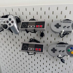 Puede incluir: Un tablero de clavijas blanco con cinco mandos de videojuegos. Los mandos son un mando Sony Playstation, dos mandos Super Nintendo y dos mandos Nintendo Entertainment System. Los mandos est&aacute;n colgados del tablero de clavijas con sus cables.