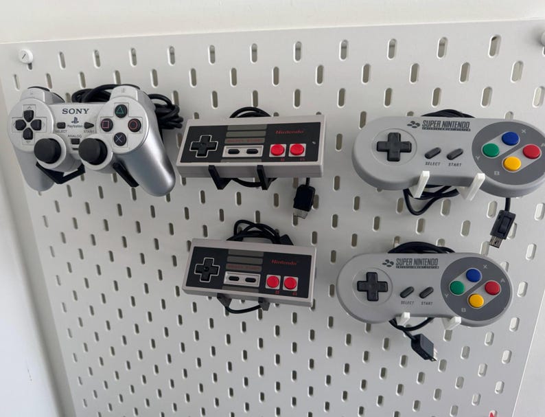 Puede incluir: Un tablero de clavijas blanco con cinco mandos de videojuegos. Los mandos son un mando Sony Playstation, dos mandos Super Nintendo y dos mandos Nintendo Entertainment System. Los mandos est&aacute;n colgados del tablero de clavijas con sus cables.
