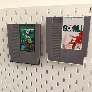 Pode incluir: Dois cartuchos de jogos cinzentos do Nintendo Entertainment System. Um apresenta um gr&aacute;fico de jogador de t&ecirc;nis com a palavra "TENNIS". O outro tem um gr&aacute;fico de jogador de futebol com a palavra "GOAL!". Ambos s&atilde;o montados numa placa perfurada branca.