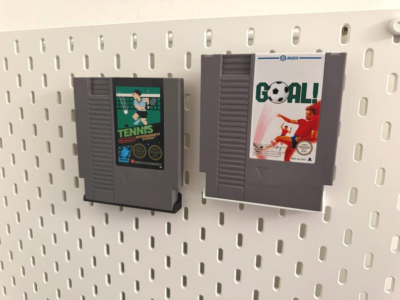 Pode incluir: Dois cartuchos de jogos cinzentos do Nintendo Entertainment System. Um apresenta um gr&aacute;fico de jogador de t&ecirc;nis com a palavra "TENNIS". O outro tem um gr&aacute;fico de jogador de futebol com a palavra "GOAL!". Ambos s&atilde;o montados numa placa perfurada branca.