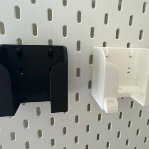 May include: Two wall-mounted storage containers, one black and one white, are shown against a white pegboard background. The containers have a curved design with a rectangular opening and are designed to hold small items.