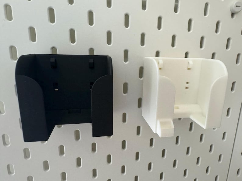 May include: Two wall-mounted storage containers, one black and one white, are shown against a white pegboard background. The containers have a curved design with a rectangular opening and are designed to hold small items.