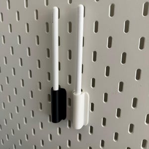 Op de afbeelding: Twee witte styluspennen worden op hun plaats gehouden door twee verticale houders, één zwarte en één witte, die zijn gemonteerd op een lichtgrijs geperforeerd bord met een herhalend patroon van verticale ovale sleuven.