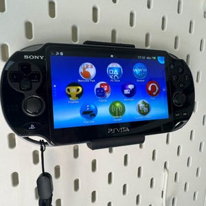 Puede incluir: Una consola de juegos portátil Sony PlayStation Vita negra con una pantalla azul que muestra el menú de inicio. La consola tiene una disposición de botones en blanco y negro y una correa negra adjunta.
