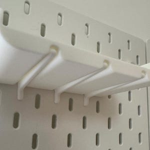 May include: White plastic wall-mounted shelf with three slots for organizing items. The shelf is mounted on a white pegboard.