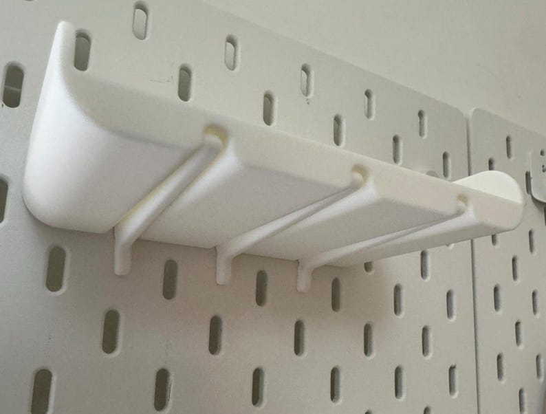 May include: White plastic wall-mounted shelf with three slots for organizing items. The shelf is mounted on a white pegboard.