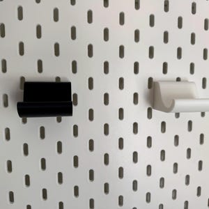 K&ouml;nnte beinhalten: Zwei Lochwandhaken, einer schwarz und einer wei&szlig;, werden vor einem wei&szlig;en Lochwandhintergrund gezeigt. Die Haken sind zur Aufbewahrung konzipiert und bestehen aus einem festen Material.