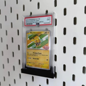 以下が含まれることがあります： 透明な保護ケースに入ったピカチュウのポケモンカード。カードは「Pikachu Berkemeja Batik」のテキストが入った黄色と緑のデザインです。カードは白い背景に黒いスタンドに表示されています。