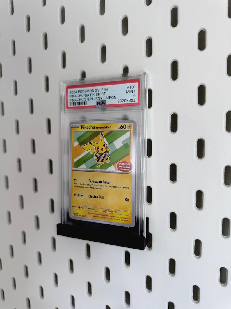 以下が含まれることがあります： 透明な保護ケースに入ったピカチュウのポケモンカード。カードは「Pikachu Berkemeja Batik」のテキストが入った黄色と緑のデザインです。カードは白い背景に黒いスタンドに表示されています。