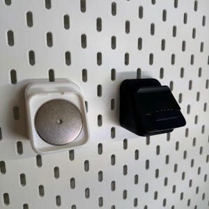 May include: Two pegboard accessories: a white magnetic holder with a silver disc and a black angled holder. Both are mounted on a white pegboard with evenly spaced oval holes. The accessories appear to be made of plastic.