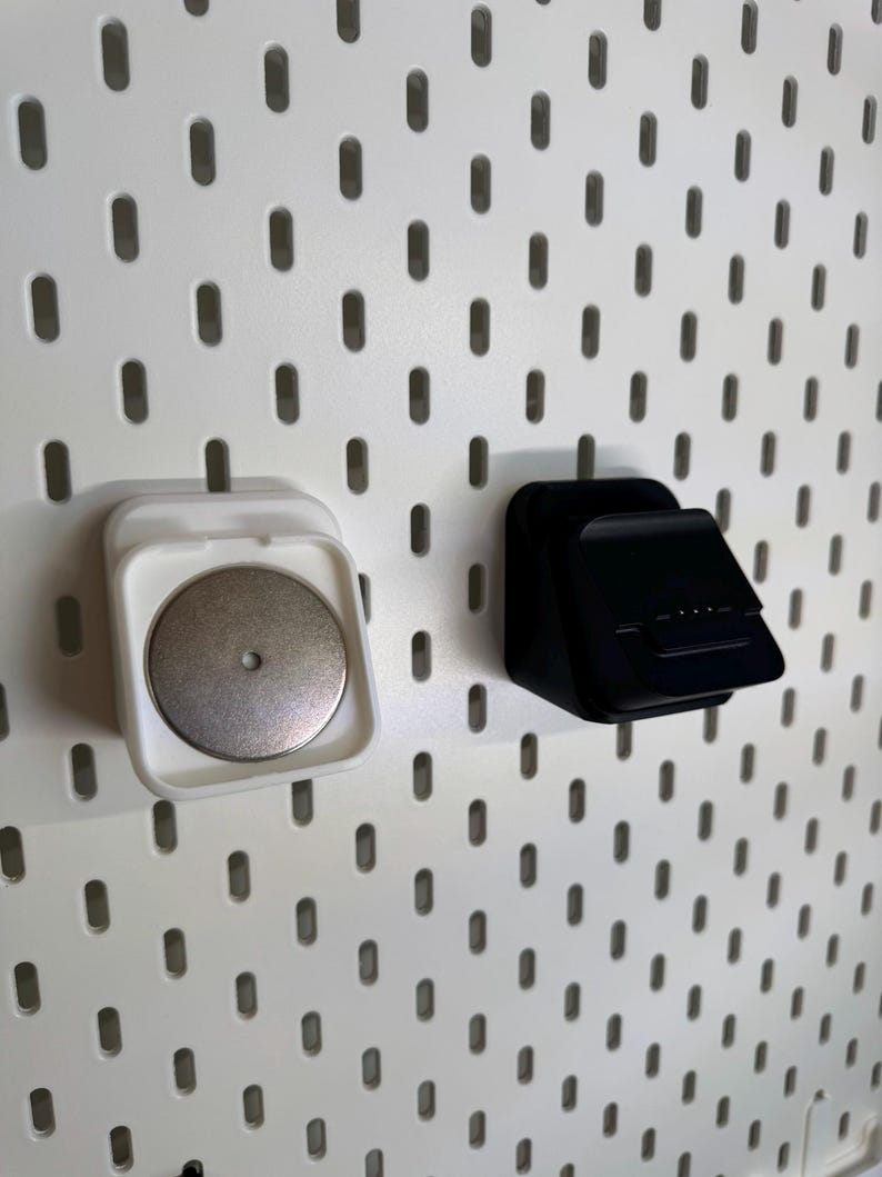 May include: Two pegboard accessories: a white magnetic holder with a silver disc and a black angled holder. Both are mounted on a white pegboard with evenly spaced oval holes. The accessories appear to be made of plastic.