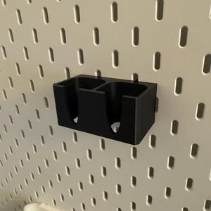 Puede incluir: Un organizador negro impreso en 3D con dos compartimentos, montado en una pared perforada blanca. El organizador es rectangular con aberturas redondeadas. La pared perforada tiene una cuadr&iacute;cula de agujeros espaciados.